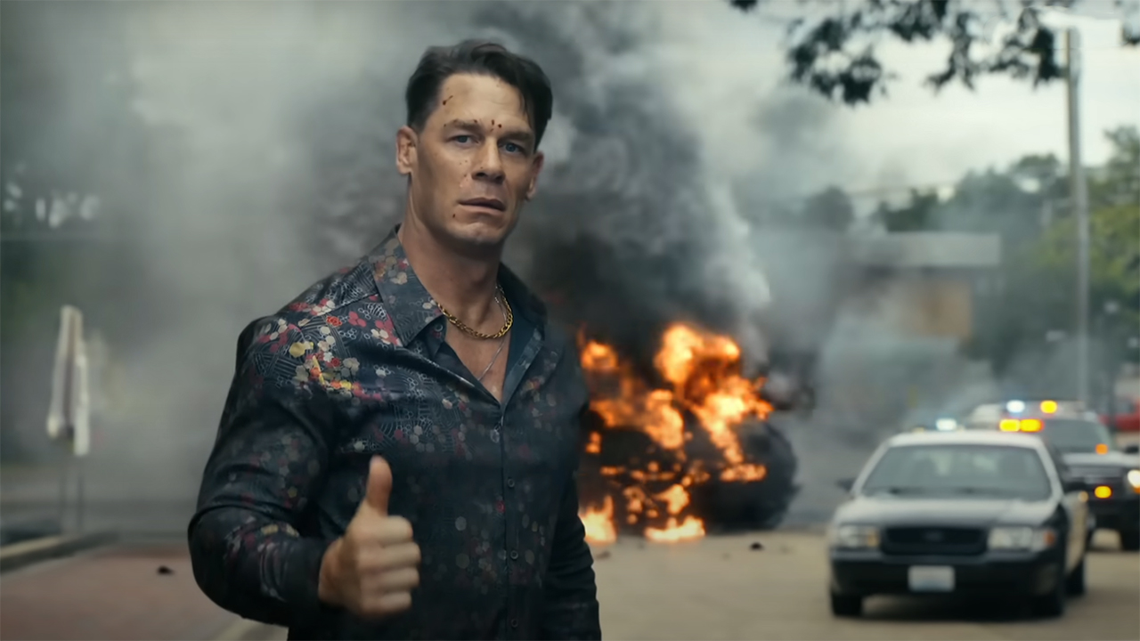 Christopher Smith giving a thumbs up with a car on fire in the background in Peacemaker season 2