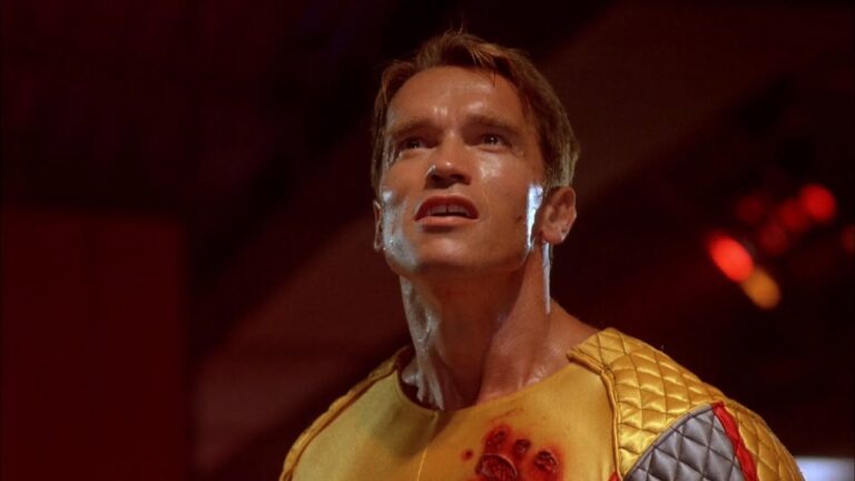 A still from the trailer for the original Running Man movie showing Arnold Schwarzenegger in a yellow form-fitting suit with blood stains